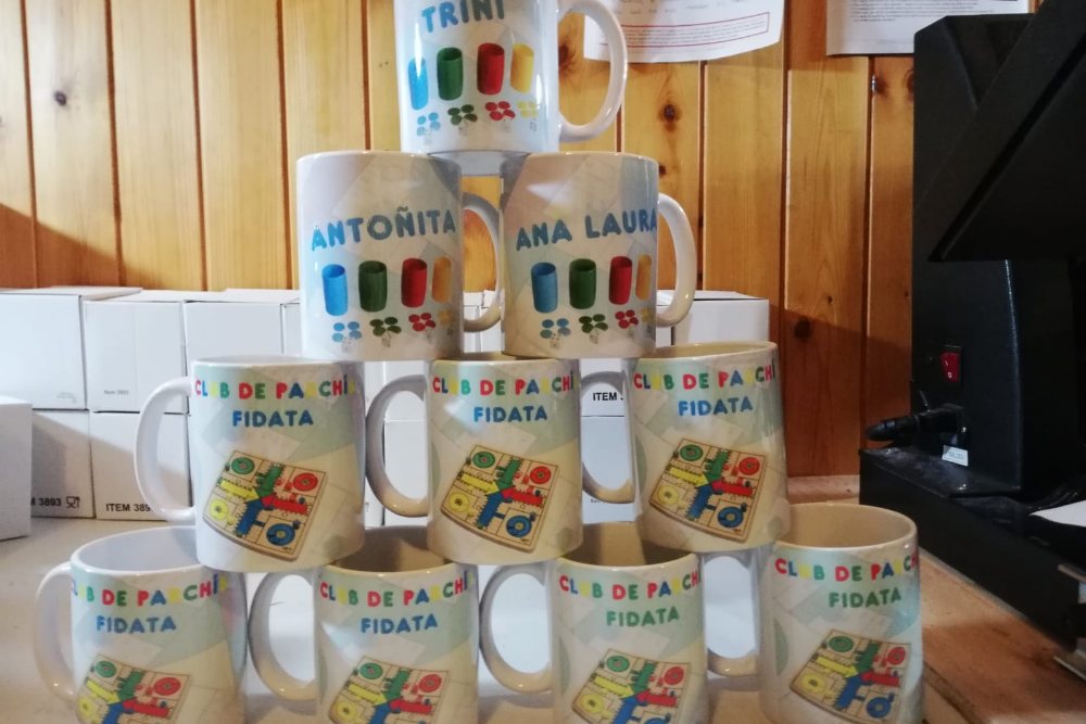 Pirámide de tazas a todo color para el Club de Parchís Fidata, cada una con el nombre de un socio, sobre la mesa del taller